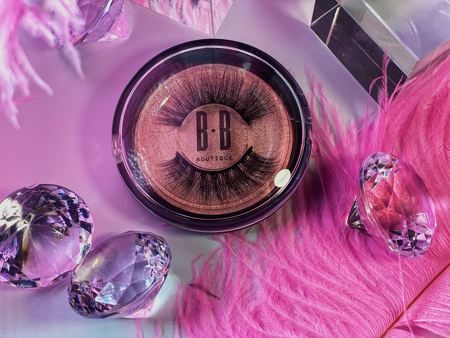 Pink image with diamonds in bottom left corner, pink ostrich feathers with glass cubes around the Foxy Lash packaging.