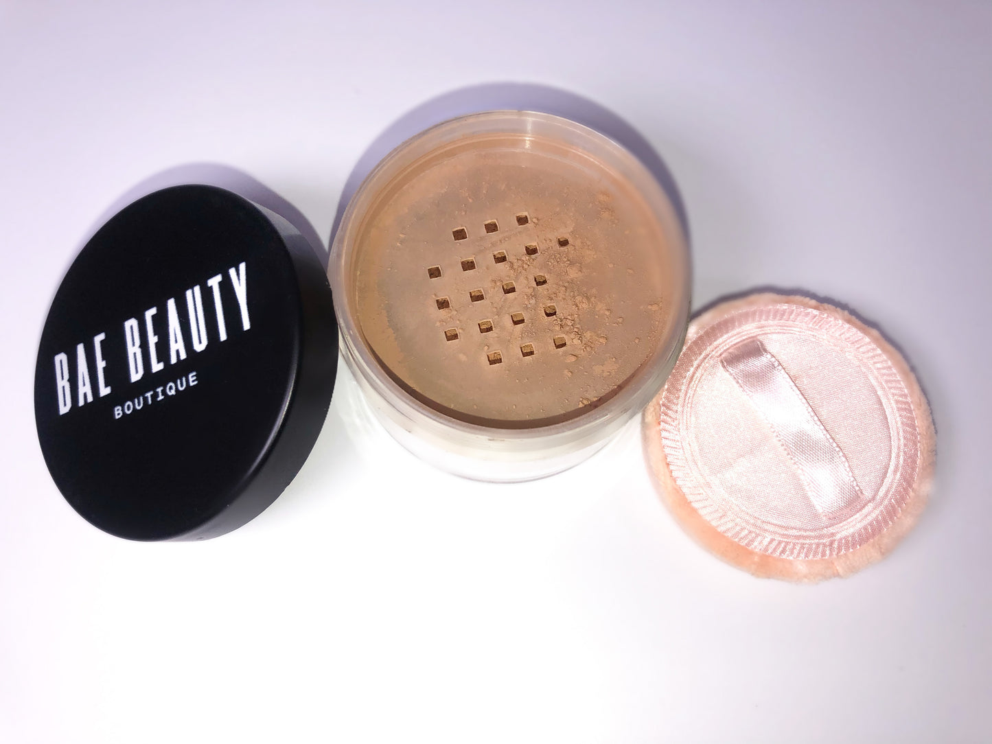 Circular container of setting powder with pink powder puff with a silk band across for holding, with lid off to the left hand side.